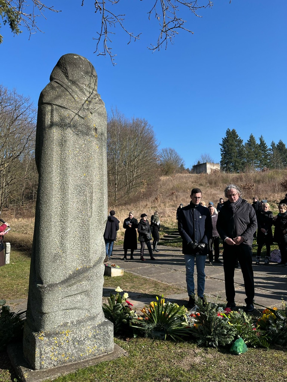 Rainer und Marcel stehen vor dem Frauenehrenmal. Vor ihnen liegen zahlreiche Blumen.